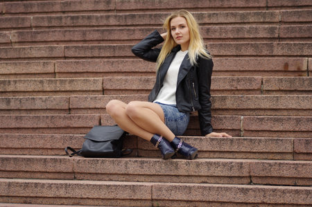 A beautiful blonde woman in a denim (jeans) skirt above the knee, white cardigan, black leather jacket, boots, sits on the steps of stone staircase. Near black bag. Street fashion, casual styleの写真素材