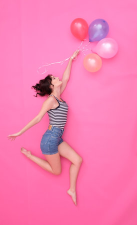 Pretty girl is jumping in the sky with baloons. Wearing jeans shorts and striped t-shirt. With fluttering long brown hair. Baloons of pastel colors.の写真素材