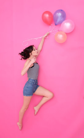 Young woman is flying on baloons. She wears jeans shorts and striped t-shirt. With fluttering long brown hair. Pink and pastel tones.の写真素材