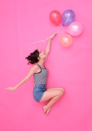 Pretty girl is jumping in the sky with baloons. Wearing jeans shorts and striped t-shirt. With fluttering long brown hair. Baloons of pastel colors.の写真素材