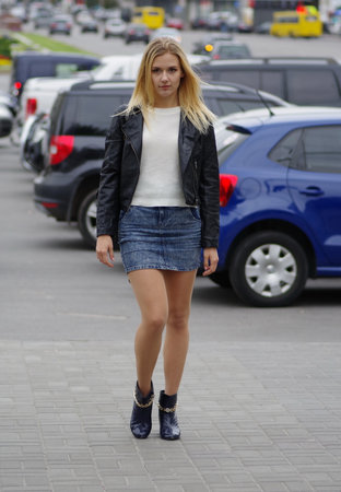 Pretty blonde girl is walking in a street, wearing short jeans skirt, white sweater and black leather coat.の写真素材