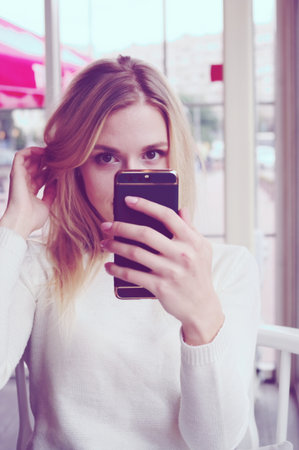 Portrait of a blonde in a white sweater making a selfie smartphone. Attentive look at the phone in his hand.の写真素材