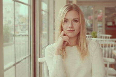 Portrait of a smiling girl dressed in a white knitted sweater in a cozy cafe. Blond hair, open smile. White chairs with a semicircular back in the background.の写真素材