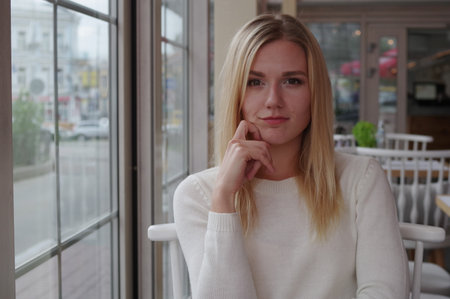 Portrait of a smiling girl dressed in a white knitted sweater in a cozy cafe. Blond hair, open smile. White chairs with a semicircular back in the background.の写真素材
