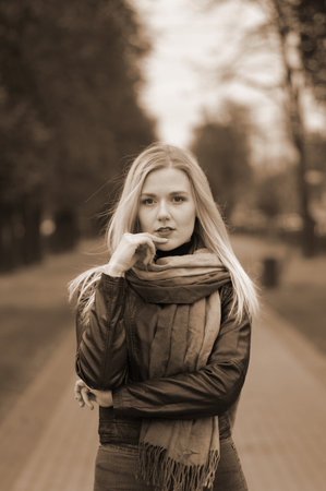 Beautiful young girl in the park on the pavement. She is standing with brooding eyes. With long blonde hair. She is wearing tight jeans, black leather jacket, scarf. Posing in autumn park.の写真素材