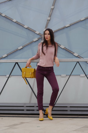 Beautiful girl leaning against a railing near a glass dome. She has in burgundy jeans, pink shirt and yellow high heels on her. With yellow handbag in her right hand. Wind plays with her brunette hair.の写真素材