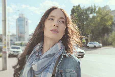 Young woman posing while walking around the city in windy weather. She wears a blue jeans jacket with a scarf. The wind plays with her beautiful long brown hair with curls.の写真素材