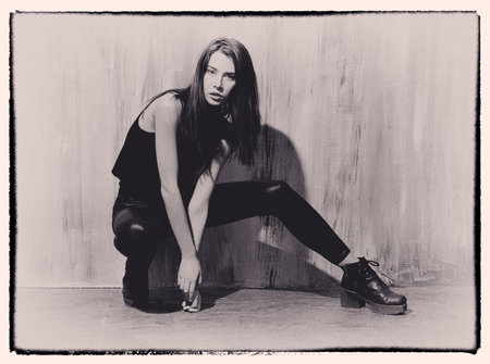Fashion portrait of dark-haired young elegant girl in black.T-shirt and leather leggings. Sitting pose: shoulders open, hands hanging between legs, laced shoes on the heel.の写真素材