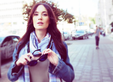 Beautiful brunette girl is walking in a city street. She is wearing a blue jeans coat and blue scarf. Holding sunglasses in her hands.の写真素材