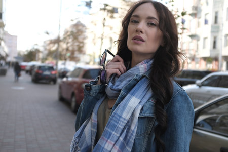 Handsome brunette girl is walking in a city street. She is wearing a blue jeans coat and blue scarf. Holding sunglasses in her right hand.の写真素材