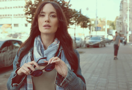 Beautiful brunette girl is walking in a city street. She is wearing a blue jeans coat and blue scarf. Holding sunglasses in her hands.の写真素材