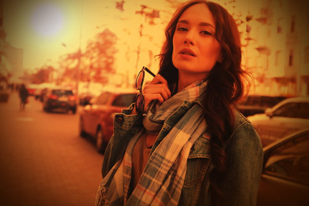 Handsome brunette girl is walking in a city street. She is wearing a blue jeans coat and blue scarf. Holding sunglasses in her right hand.の写真素材