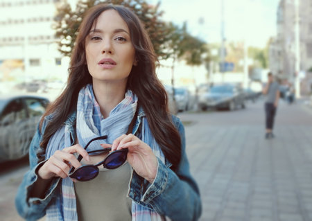 Beautiful brunette girl is walking in a city street. She is wearing a blue jeans coat and blue scarf. Holding sunglasses in her hands.の写真素材