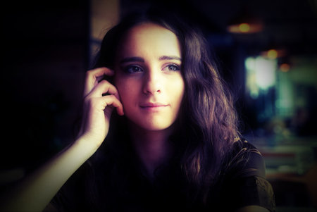 Portrait of a young girl in a brown blouse in a cafe. A beautiful girl sits in a coffee shop and looks out the window, thinking, raising her hand to her face. Big brown eyes, curly hairの写真素材