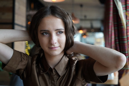 Portrait of a young girl in a brown blouse in a cafe. A beautiful girl is sitting in a cafe holding hands behind her head. Big brown eyes, curly hair, natural make-up.の写真素材
