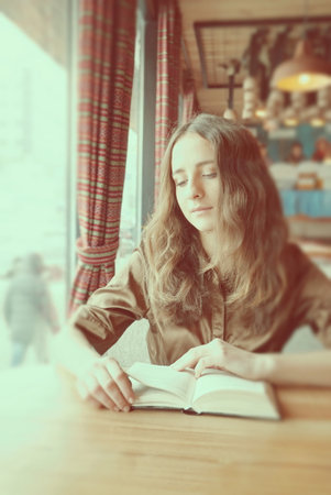 A beautiful student girl is sitting in a cafe next to the window. She is reading a book to prepare for her education syllabus. She has nice long brown hair. Wearing a brown short sleeve blouse.の写真素材