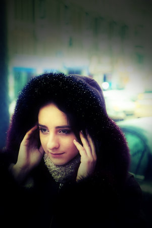 A handsome girl is standing in a street. It is winter time and it is snowing. She is wearing a coat with a hood and a scarf. Trying to hide her hair under the hood. She has long brown hair.の写真素材