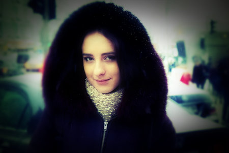 A beautiful girl is walking down a street. It is winter time and it is snowing. She is wearing a coat with a hood and a scarf, looking toward you. She has long brunette hair. Snowflakes on her hood.の写真素材