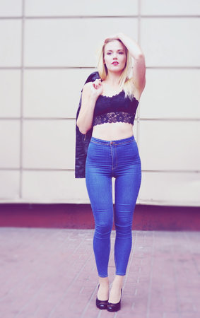 Sexy pretty girl is posing on a street. Wearing blue jeans and black crop top. Her leather coat is in her right hand on the shoulder.の写真素材