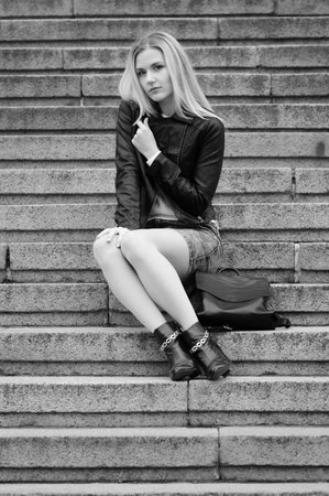 Pretty young girl is sitting on a marble stairs. Her handbag is near her legs. Wearing jeans skirt, black leather coat. She has nice long blonde hair.の写真素材