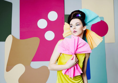 A female model in yellow is actively posing in the studio on an unusual background. Fashion story, Danish design. Color composition. Eccentric attire. Short dress, pink bow of paper in hand.の写真素材