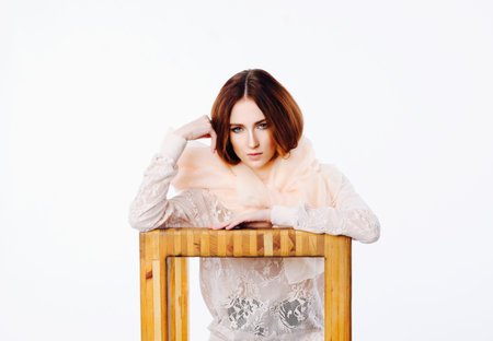 The red-haired girl is playfully posing in a lace blouse and a translucent scarf sitting on a wooden chair. . Her legs are bent at the knees, Hands - on the elbows. Grace, thoughtfulnessの写真素材
