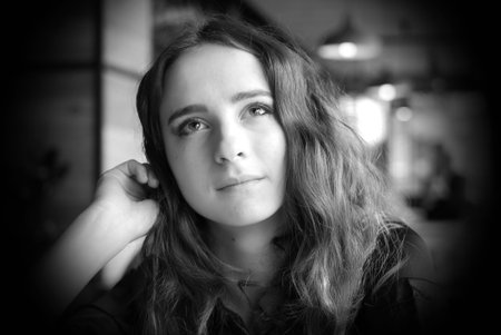 Portrait of a young girl in a brown blouse in a cafe. A beautiful girl sits in a coffee shop and looks out the window, thinking, raising her hand to her face.の写真素材