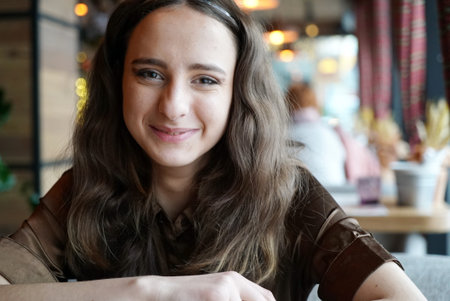Portrait of a young girl in a brown blouse in a cafe. Beautiful girl is sitting in a cafe. Big brown eyes, curly hair, natural make-up. A smile on her face.の写真素材