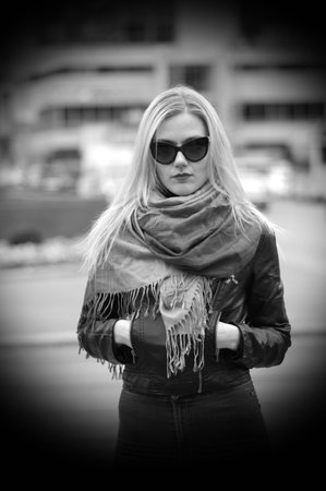 Serious young female is standing in a city street. Wearing blue jeans, leather jacket, rose scarf and dark sunglasses. Cars and buildings are on the background.の写真素材