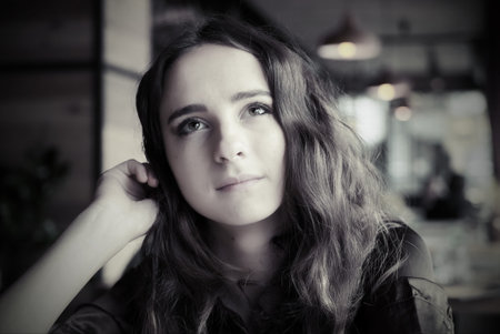 Portrait of a young girl in a brown blouse in a cafe. A beautiful girl sits in a coffee shop and looks out the window, thinking, raising her hand to her face.の写真素材