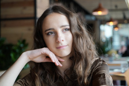 Portrait of a young girl in a brown blouse in a cafe. A beautiful girl sits in a coffee shop and looks out the window, thinking, raising her hand to her face.の写真素材