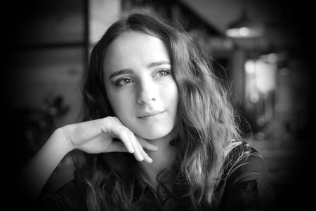 Portrait of a young girl in a brown blouse in a cafe. A beautiful girl sits in a coffee shop and looks out the window, thinking, raising her hand to her face.の写真素材