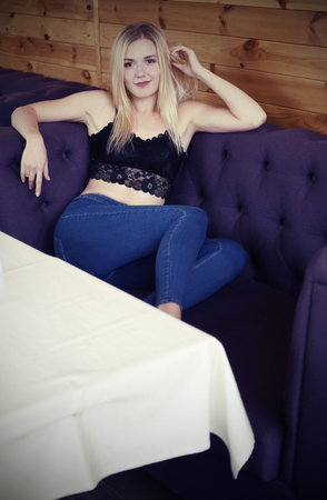 Portrait of a blonde girl in a black lace crocheted top and jeans, sitting in a playful pose on the couchの写真素材