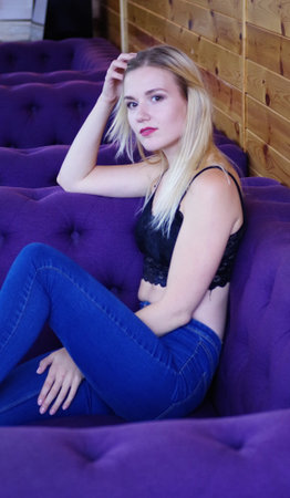 Portrait of a blonde girl in a black lace crocheted top and jeans, sitting in a playful pose on the couch.の写真素材