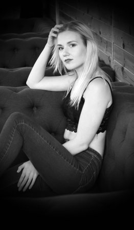 Portrait of a blonde girl in a black lace crocheted top and jeans, sitting in a playful pose on the couch.の写真素材