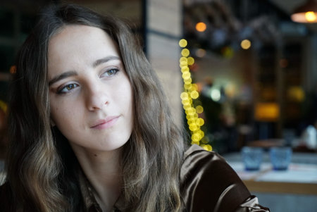Lonely pretty girl is sitting in the cafe. She has big brown eyes and nice brown long hair. Wearing a brown silk blouse. With light flares on the background.の写真素材
