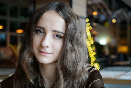 Handsome girl is sitting in the cafe. She has big brown romantic eyes. With nice brown long hair. Wearing a brown silk blouse. With light flares on the background.の写真素材