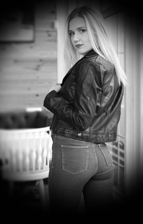Beautiful young blonde woman posing in cafe. She wears a black leather jacket and blue jeans. Large bright windows in the cafe. Sexual pose, jeans, backsideの写真素材