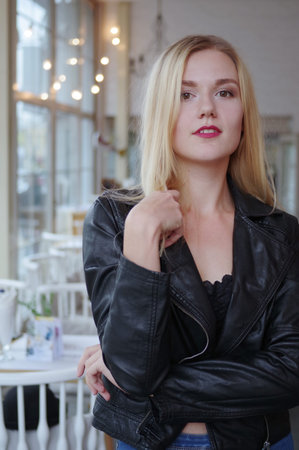 Beautiful young blonde woman posing in cozy cafe. She wears a black leather jacket and blue jeans. Large bright windows in the cafeの写真素材