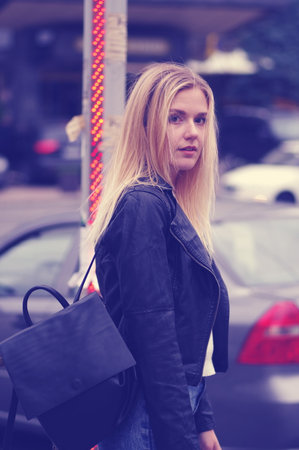 Pretty blonde girl posing outdoors. The girl is dressed in a black leather jacket, a white sweater and a denim skirt. On his shoulder is a black bag.の写真素材
