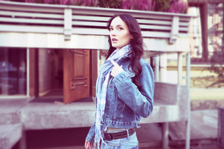 Beautiful young woman is walking and posing outdoors. She has beautiful long brown hair with curls. She wears a blue jeans jacket with a scarf.の写真素材