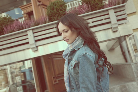 Beautiful young woman is walking and posing outdoors. She has beautiful long brown hair with curls. She wears a blue jeans jacket with a scarf.の写真素材