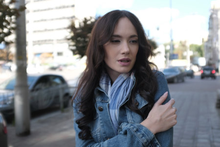 A beautiful young woman is walking and posing somewhere in the city. She has nice long brown hair with curls. She is wearing blue jeans jacket with scarf.の写真素材
