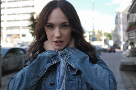 A beautiful young woman is walking and posing somewhere in the city. She has nice long brown hair with curls. She is wearing blue jeans jacket with scarf.の写真素材