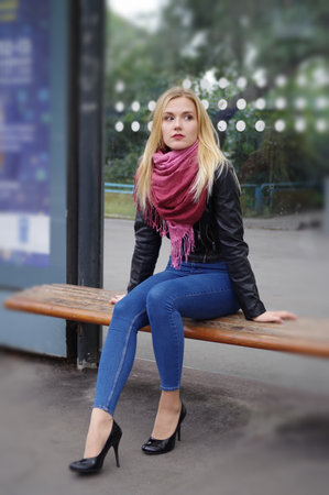 A beautiful blonde girl is sitting at a public transport stop. The girl is dressed in a black jacket and blue jeans with a high waist. A voluminous shawl of pink color is tied around neckの写真素材