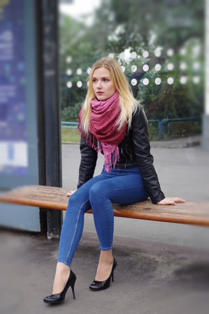 A beautiful blonde girl is sitting at a public transport stop. The girl is dressed in a black jacket and blue jeans with a high waist. A voluminous shawl of pink colorの写真素材