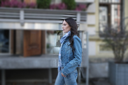 A beautiful young woman is walking and posing against flower shop in the city. She has nice long brown hair with curls. She is wearing blue jeans jacket with scarf.の写真素材
