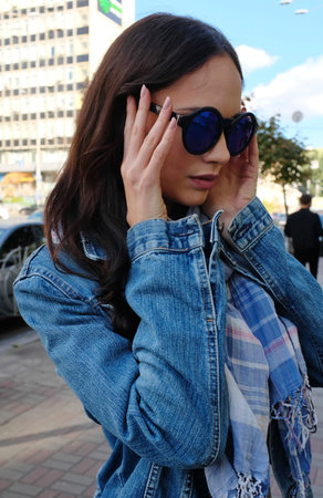 Beautiful young woman is walking and posing somewhere in the city. She wears a blue jeans jacket with a scarf. Large sunglasses.の写真素材