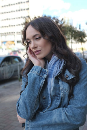 A beautiful young woman is walking and posing somewhere in the city. She has nice long brown hair with curls. She is wearing blue jeans jacket with scarf.の写真素材