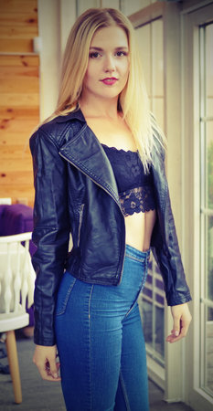 Beautiful young blonde woman posing in cozy cafe. She wears a black leather jacket, black lacy tops and blue jeans. Large bright windows in the cafe. Sexual posture.の写真素材
